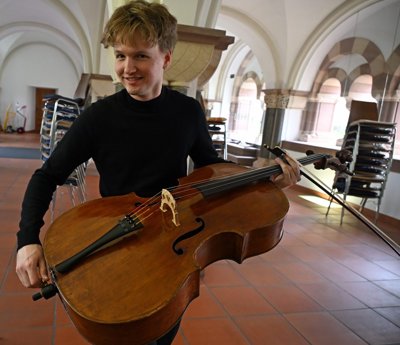Carlo Lay steht lächelnd mit Cello und Bogen im Kreuzgang des Klosters