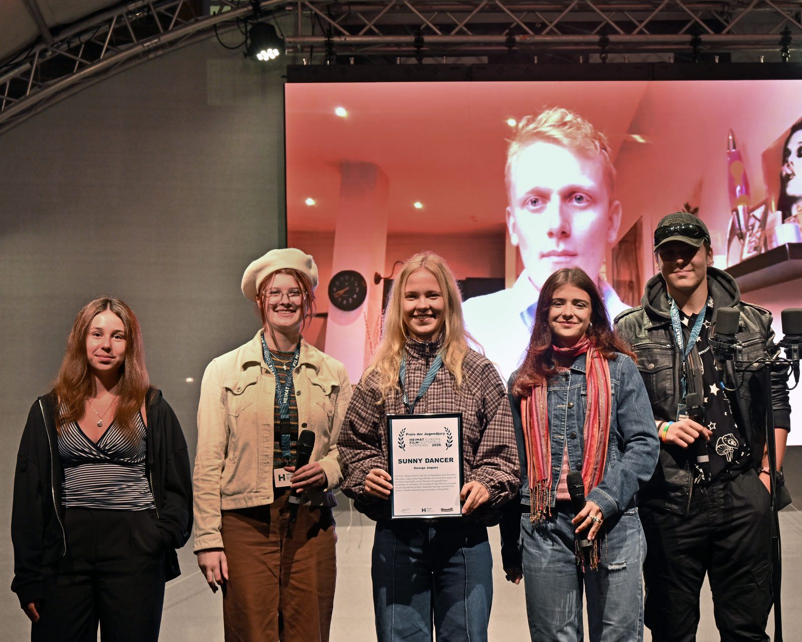 Die fünf Mitglieder der Jugendjury stehen mit der Urkunde für SUNNY DANCER auf der Bühne, im Hintergrund der zugeschaltete Regisseur George Jaques